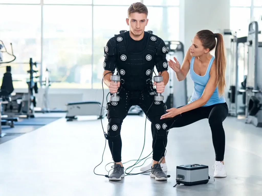 Mężczyzna w kombinezonie EMS ćwiczy z hantlami pod okiem trenerki w nowoczesnym, jasnym studiu fitness.