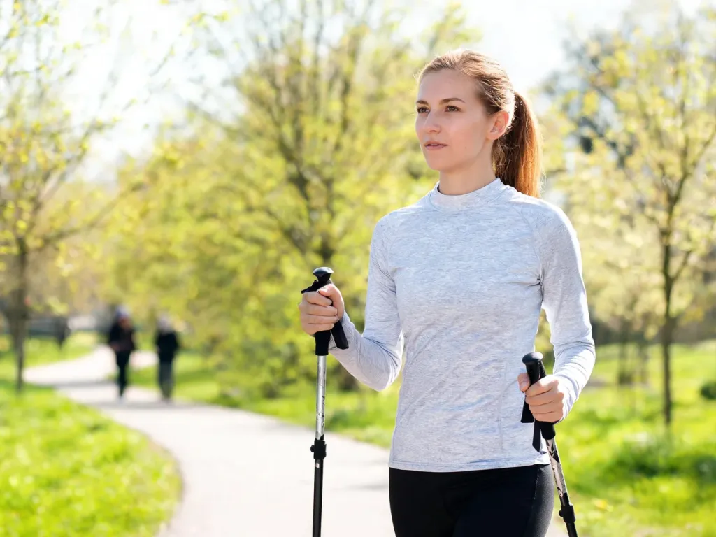 Kobieta w sportowym stroju maszeruje z kijami nordic walking w słonecznym parku, prezentując prawidłową technikę kroku.